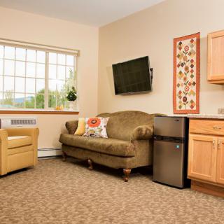 A cozy living room with a couch, armchair, and a mounted television, complemented by wall art and a mini fridge.