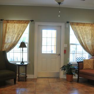 A cozy living room with a central door, flanked by windows adorned with curtains, and featuring comfortable seating and decor.