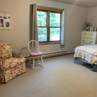A simple and cozy bedroom featuring a floral chair, a white dresser, and ample natural light coming through the window.