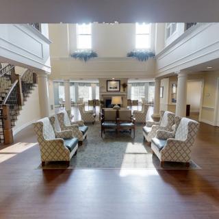 A spacious living room with elegant furniture, featuring multiple chairs and sofas arranged around a central area rug, with large windows allowing natural light to fill the space.
