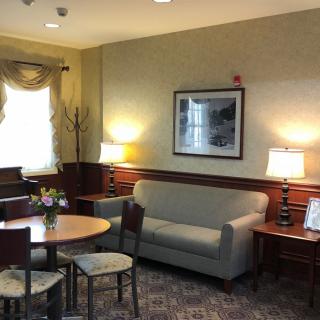 A cozy living room with a sofa, table, chairs, and lamps, featuring artwork on the wall and a window with drapes.
