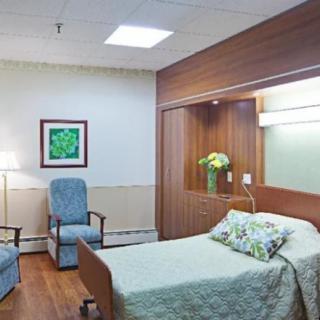 A clean and well-organized hospital room featuring a bed, chairs, artwork, and a vase of flowers.