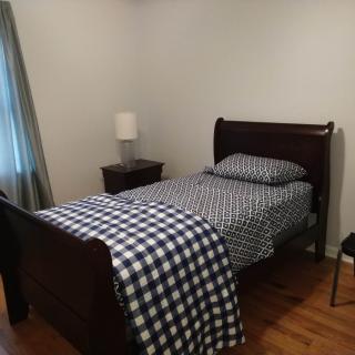 A simple bedroom with a single bed featuring checkered bedding, a nightstand with a lamp, and hardwood flooring.