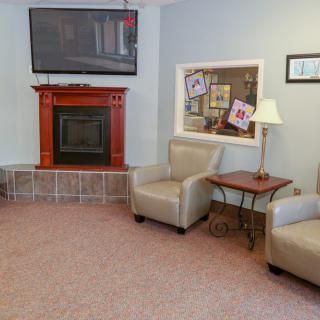 A cozy living room with a fireplace, two armchairs, a table, and a wall-mounted television.