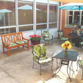 A sunny patio area featuring tables, chairs, a bench, umbrellas, and vibrant flowers.