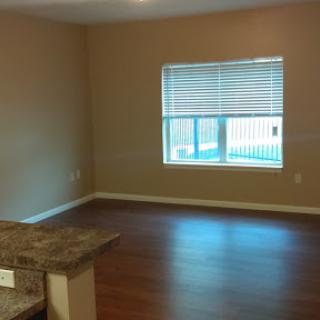 A spacious living area featuring natural light from a window with blinds, complemented by hardwood flooring and neutral wall tones.