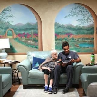 An elderly woman enjoys reading with her caregiver in a cozy, colorful indoor setting.
