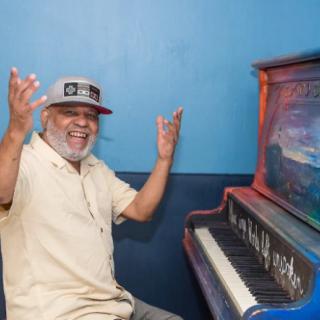 A joyful man wearing a hat sits beside a painted piano, raising his arms in excitement.