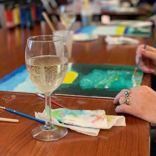 A person painting in an art class with a glass of wine nearby.