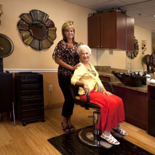 An elderly woman sits in a salon chair, smiling as a hairdresser stands behind her.