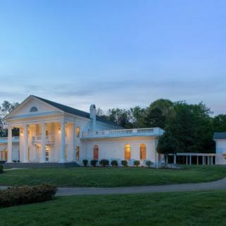 A stately mansion illuminated during the evening, surrounded by a well-kept lawn and trees.