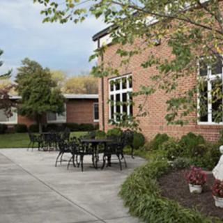 An outdoor patio area with tables and chairs next to a brick building, surrounded by trees and landscaping.