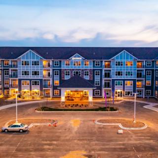 A large residential building with lit windows at dusk, featuring a spacious parking lot.