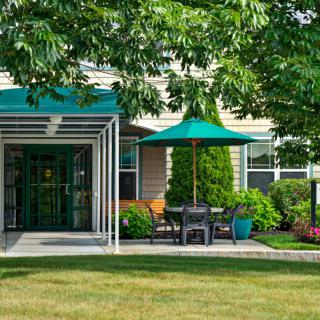 A cozy outdoor seating area with a green umbrella and canopy, surrounded by lush greenery in front of a building.