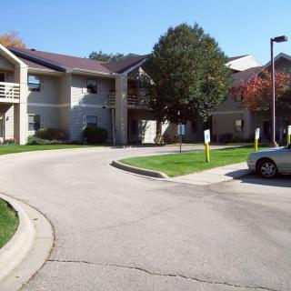 A residential apartment complex with a parking lot, surrounded by trees and cars.