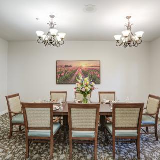 A well-lit dining room with a large table, chairs, chandeliers, and floral centerpiece beneath a scenic wall art.