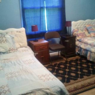A cozy bedroom featuring two beds with floral bedding, a nightstand with red lamps, a chair, and a patterned rug.