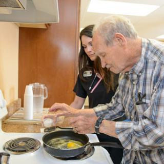 An elderly man receives assistance while cooking eggs in a kitchen setting.