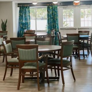 An empty dining area with multiple tables and chairs, featuring wooden floors and large windows with teal curtains.
