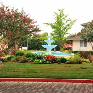A picturesque garden featuring a multi-tiered fountain surrounded by greenery and vibrant flowers, with a house in the background.