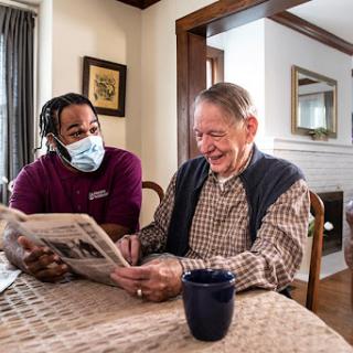 A caregiver and an elderly man share a joyful moment while looking at a newspaper together.