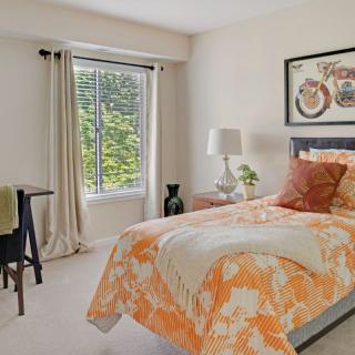 A cozy bedroom with a floral orange comforter, a bedside lamp, and a window allowing natural light.