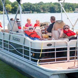 A group of people wearing life jackets relax on a pontoon boat on the lake during a sunny day.