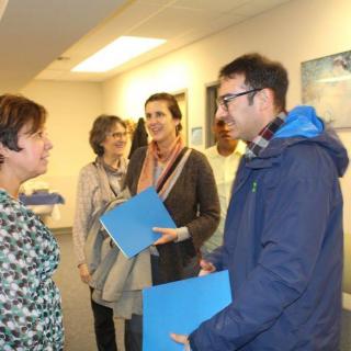 A group of people engaged in a conversation in an office setting, holding blue folders.