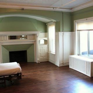 A spacious, empty room featuring green walls and a wooden floor, highlighted by natural light from the large windows.