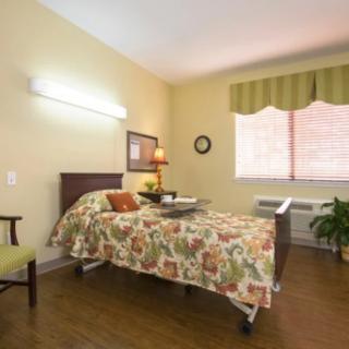 A well-lit hospital or nursing home room featuring a single bed with floral bedding, a chair, and a window with curtains.