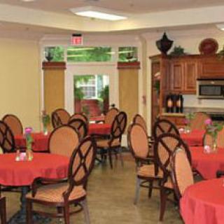 A cozy dining area with round tables covered in red tablecloths and elegant wooden chairs, featuring a kitchenette in the background.
