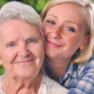 A young woman embraces an elderly woman, both smiling warmly outdoors.