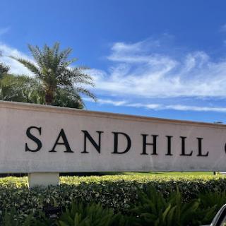 A view of the Sandhill sign against a clear blue sky, surrounded by lush greenery and palm trees.