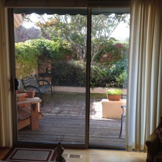 A view of a garden patio through sliding glass doors with outdoor seating and lush greenery.
