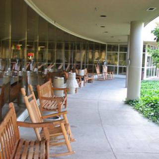 A row of wooden rocking chairs placed along a curved outdoor walkway beside large windows.