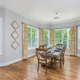 A bright dining room with hardwood floors and a table set by large windows adorned with patterned curtains.