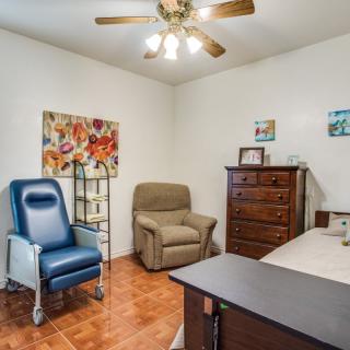 A cozy office space with a blue chair, recliner, desk, and dresser, accented by colorful artwork and a ceiling fan.