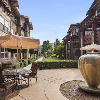 A peaceful apartment complex courtyard featuring a fountain, outdoor seating with tables and umbrellas, surrounded by greenery.