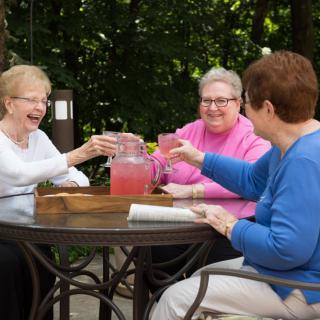 Three elderly women enjoying drinks and laughter together outdoors.
