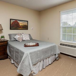A cozy bedroom with a neatly made bed, bedside table, lamp, wall artwork, and a window with blinds.