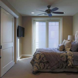 A cozy bedroom with a large bed and ceiling fan, featuring a wall-mounted television and ample natural light from the windows.