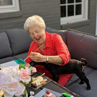 An elderly woman laughing while enjoying a meal outdoors with a small dog on her lap.