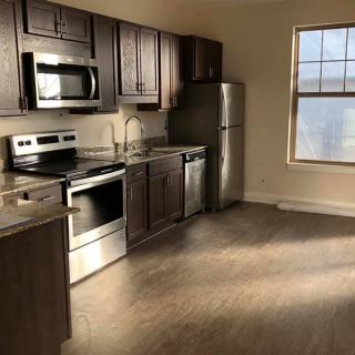 A modern apartment kitchen with stainless steel appliances and wooden cabinets, illuminated by sunlight.