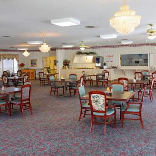 A spacious dining room with multiple round tables and chairs, featuring chandeliers and carpeted flooring.