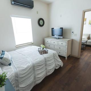 A minimalist bedroom with a white bed, dresser, and a TV, featuring wood flooring and natural light.