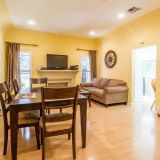 A cozy living and dining area with warm yellow walls, featuring a sofa, dining table with chairs, a TV on a fireplace mantel, and wooden flooring.