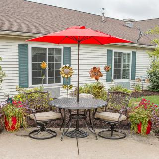 A cozy patio setting featuring a round table with four chairs and a vibrant red umbrella, surrounded by potted plants in front of a house.