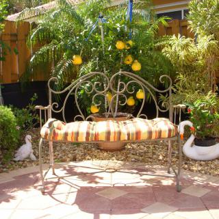 A decorative garden bench with a striped cushion, surrounded by lush plants and sunlight.