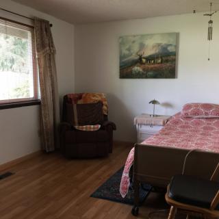 A cozy bedroom with a bed, chair, and painting on the wall, featuring wooden flooring and a window with curtains.