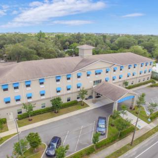 Aerial view of a hotel building with a parking lot and surrounding trees under a clear blue sky.
